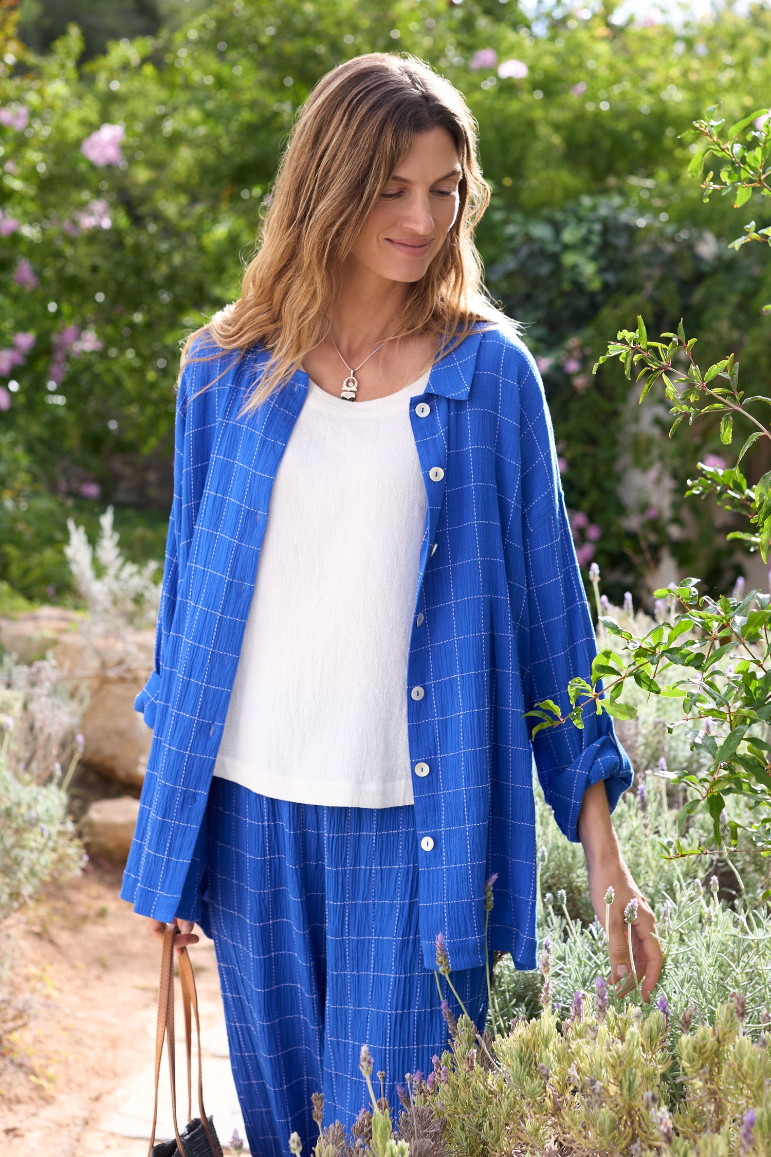 Amidst a lush garden path, a person in a relaxed silhouette showcases the classic button-through Stitched Grid Check Shirt in blue.