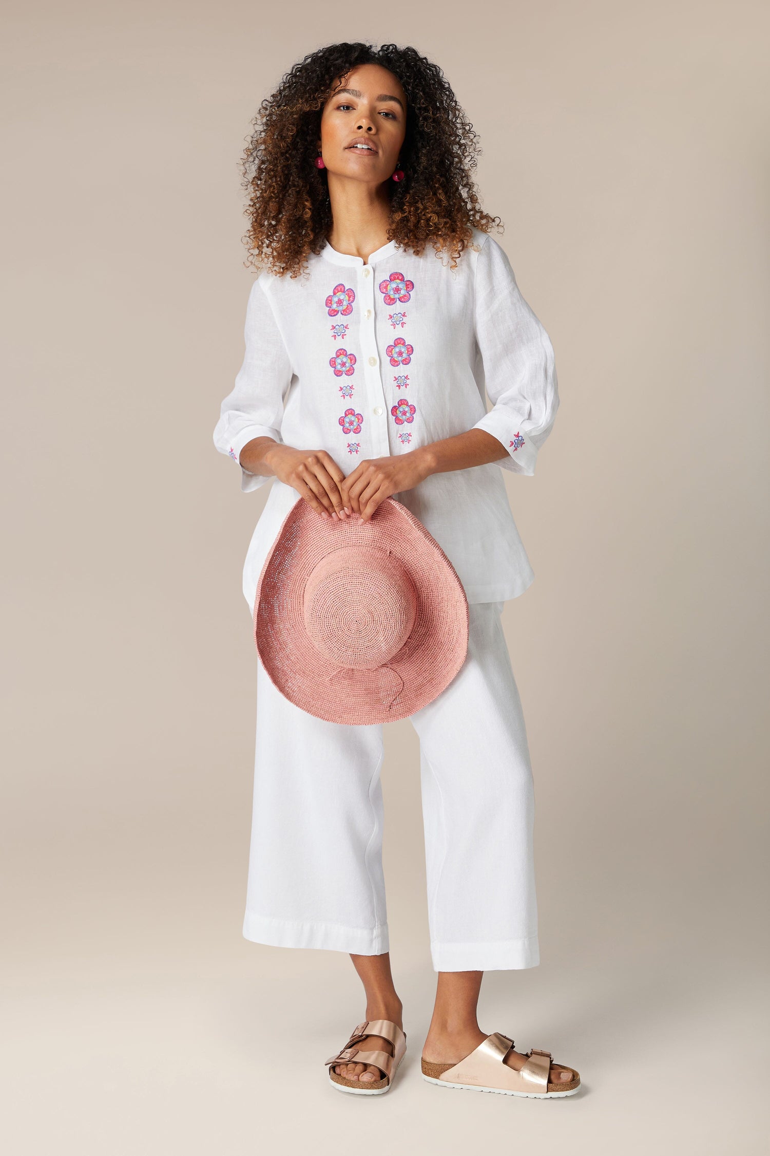 A woman stands holding a pink hat, wearing a Floral Embroidery Shirt with bracelet-length sleeves, white pants, and sandals against a neutral background.