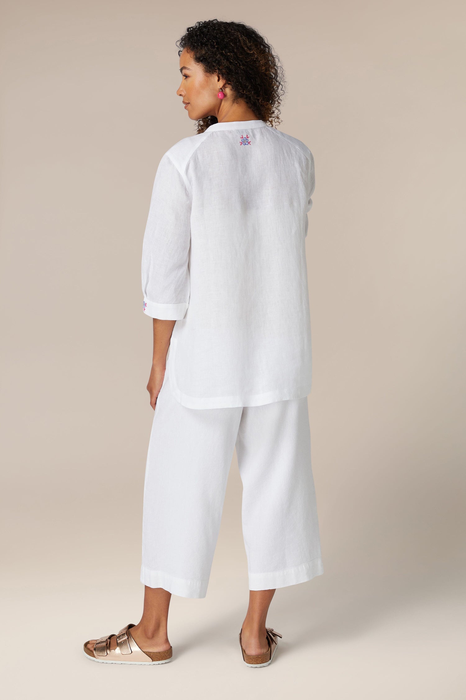 Woman viewed from the back, wearing a white Floral Embroidery shirt and cropped pants, standing against a neutral background.