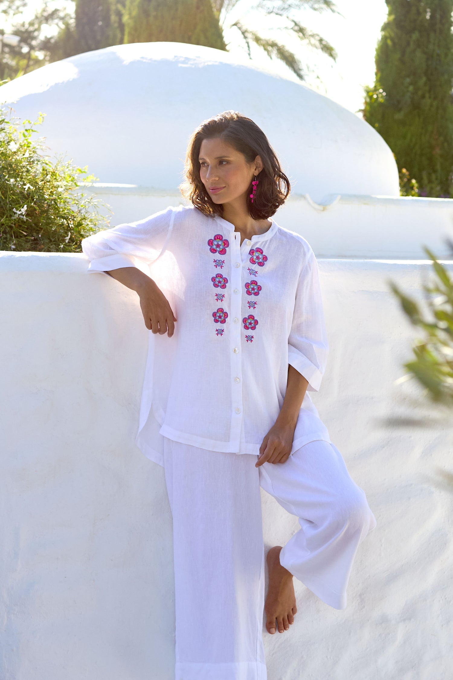 A woman in a white Rose Garden Shirt and pants stands against a white wall, lifting one leg slightly, with a sunlit garden background.