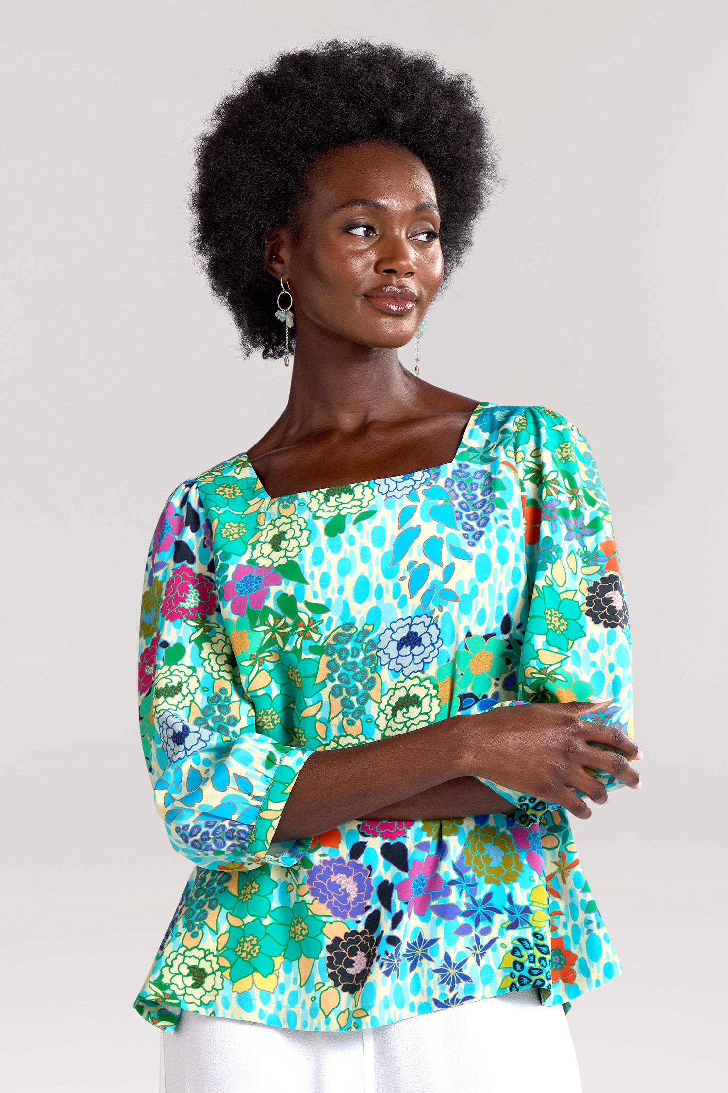 A woman with an afro hairstyle dons a colorful Garden Spot Square Neck Top with long sleeves made of draping viscose fabric and dangling earrings, posing with her arms crossed against a plain background.