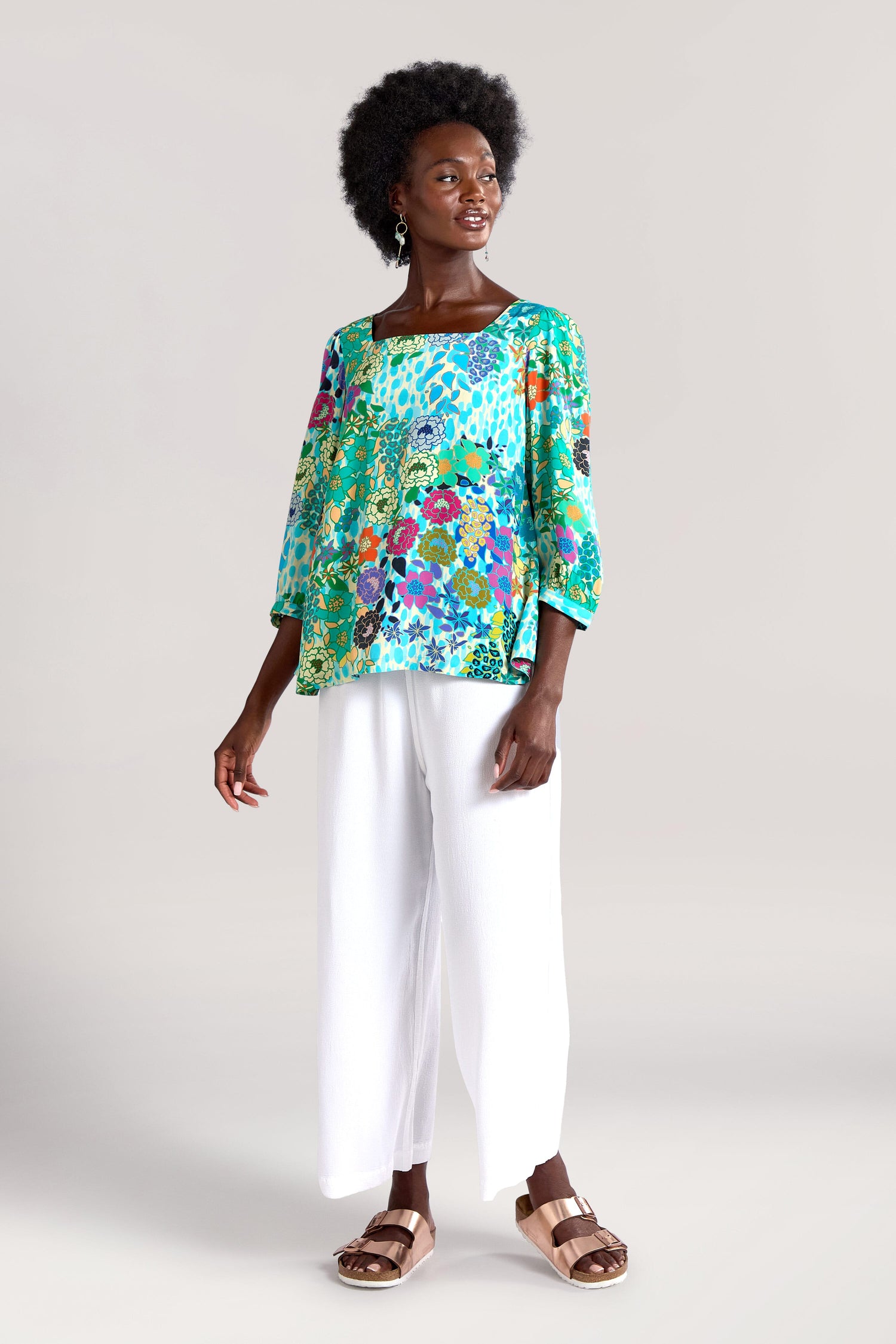 Person wearing a vibrant Garden Spot Square Neck Top made of draping viscose fabric, paired with white wide-leg pants and brown sandals, standing against a plain background.