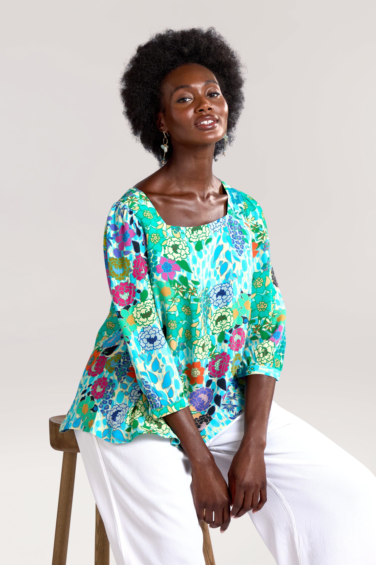 A woman with curly hair is seated on a wooden stool, wearing a colorful Garden Spot Square Neck Top and white pants. She is looking at the camera with a slight smile.