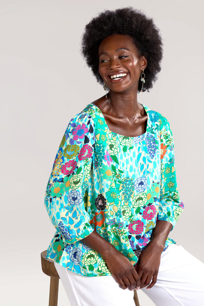 A person with an afro hairstyle smiling, wearing a Garden Spot Square Neck Top made of draping viscose fabric and white pants, seated on a wooden stool against a plain background.