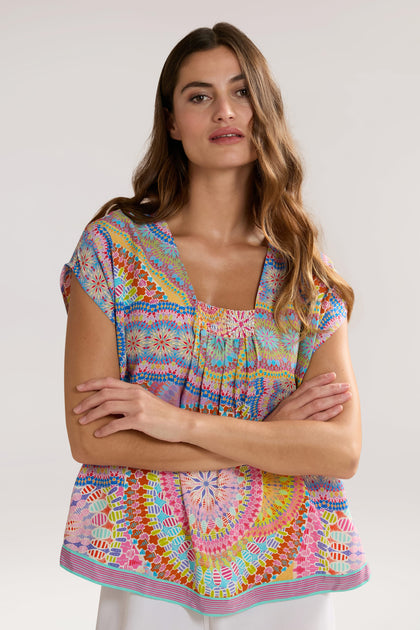 A woman with long hair stands with her arms crossed, wearing a Circle Mandala Print Top. The background is plain.