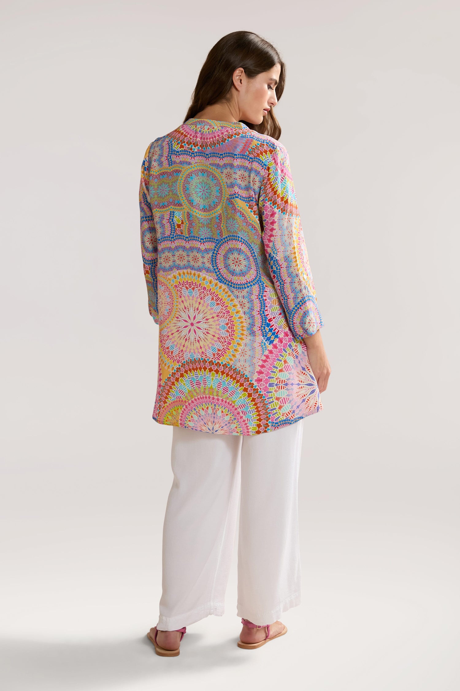 A person with long brown hair wearing a colorful, patterned tunic and white pants is standing and facing away. The Circle Mandala Print Shirt, made of pure viscose, adds an artistic flair to the ensemble.