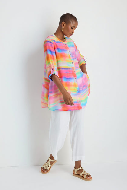 A person stands against a white wall wearing the Painted Horizon Flare Shirt, white pants, and beige sandals.