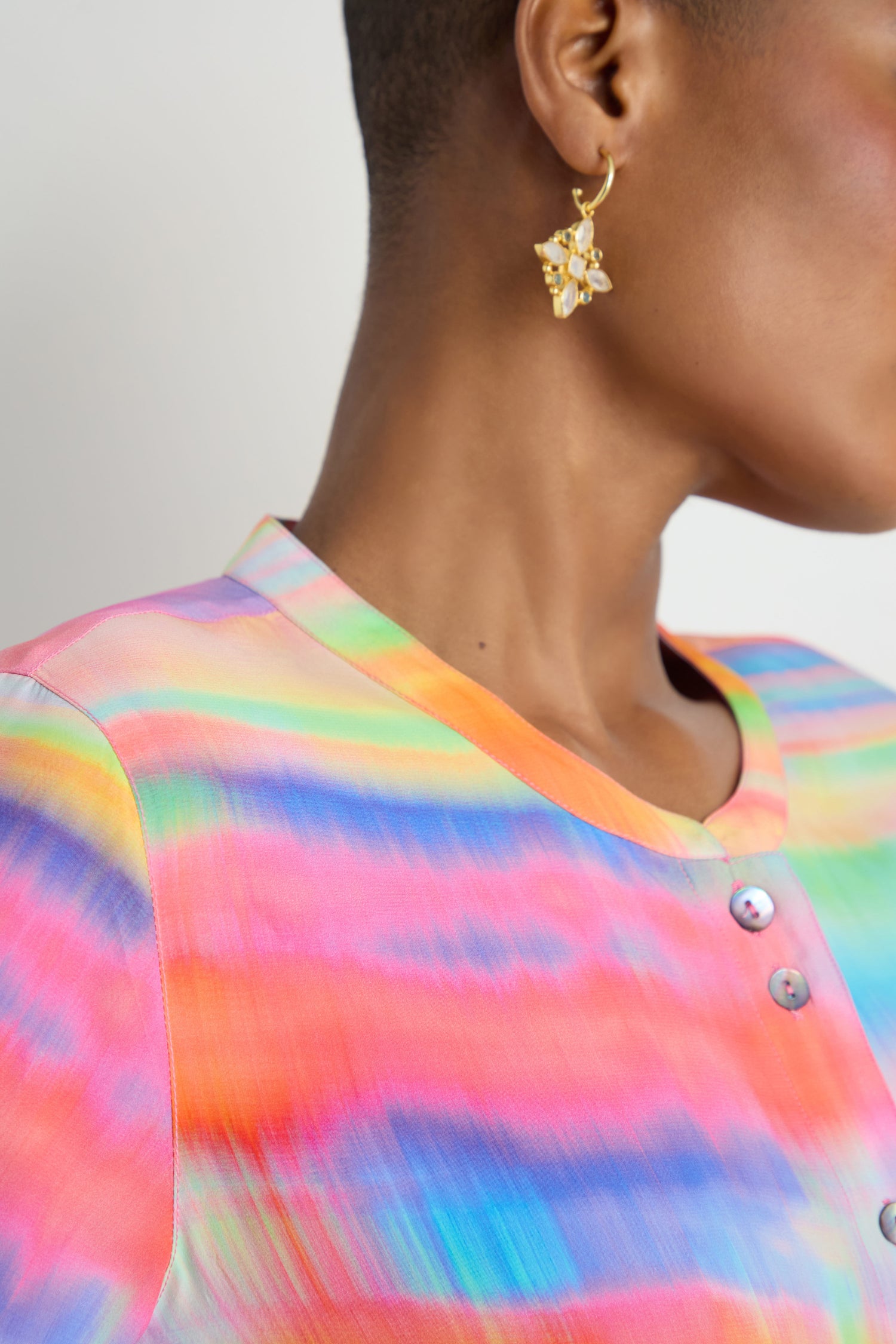Close-up of a person wearing the Painted Horizon Flare Shirt, featuring a colorful rainbow pattern and buttons, paired with a gold floral earring.