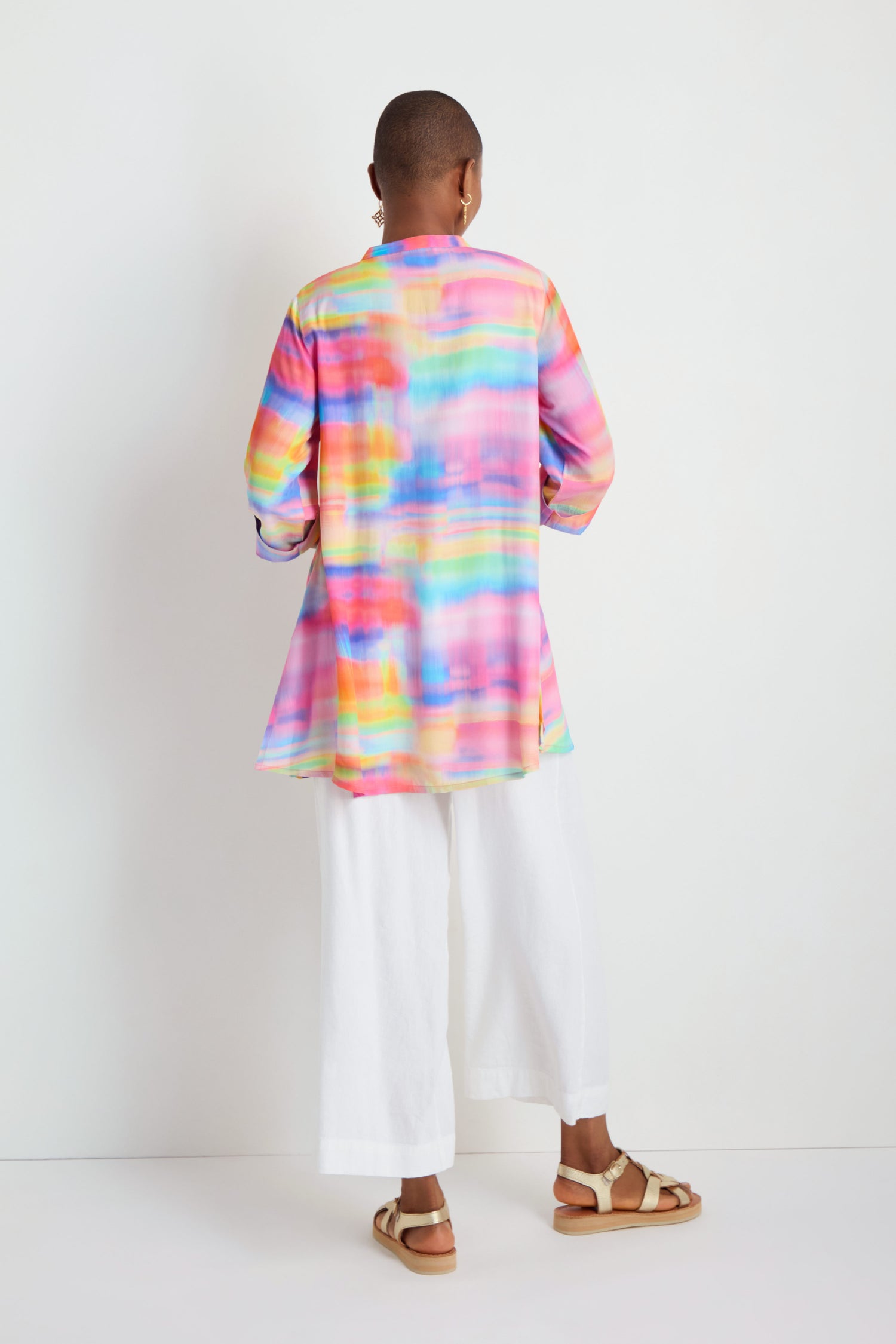 A person stands facing away, wearing the Painted Horizon Flare Shirt—a relaxed fit, colorful long-sleeved tunic with a watercolor pattern—paired with white wide-leg pants and tan sandals against a plain background.
