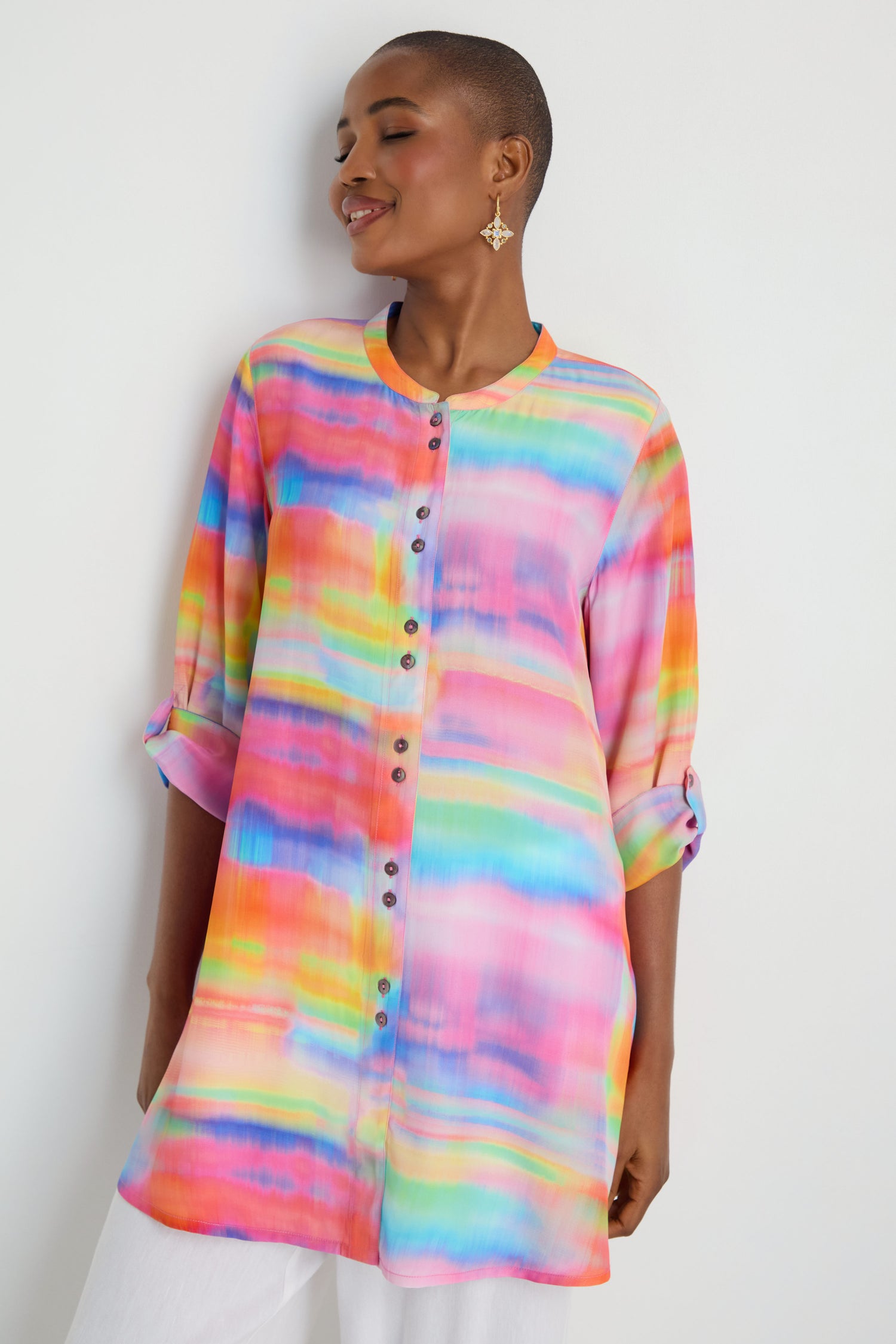 A person models the Painted Horizon Flare Shirt, a colorful long-sleeve button-up tunic with a Nehru collar and watercolor pattern, rolled cuffs, paired with white pants against a plain white background.