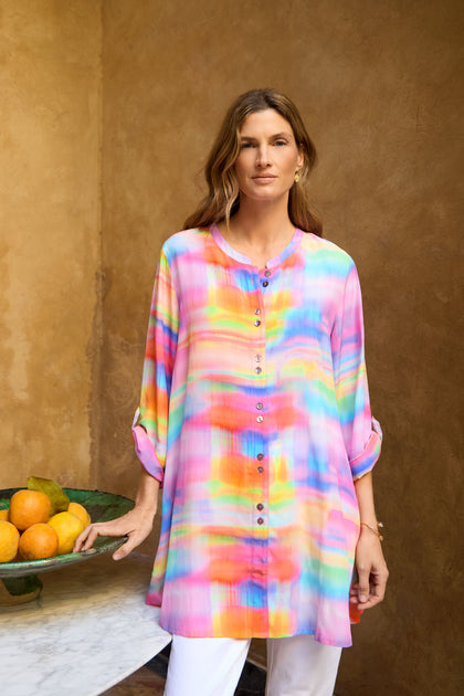 A woman indoors by a marble table holds a bowl of oranges while wearing the Painted Horizon Flare Shirt, style GRT5835-PH—a colorful, loose-fitting, button-up tunic with a watercolor pattern.