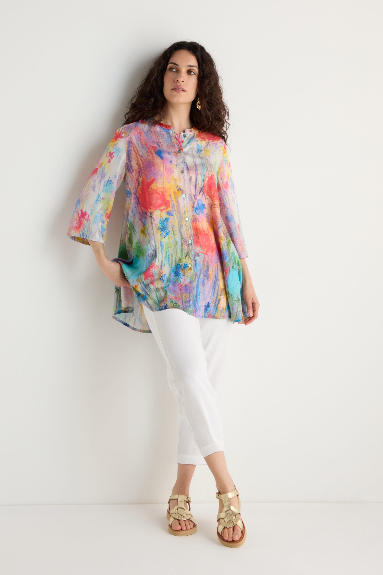 A woman stands against a white wall wearing the Meadow Dreams Flared Shirt, featuring a colorful watercolor floral print and loose fit, paired with white pants and beige sandals.