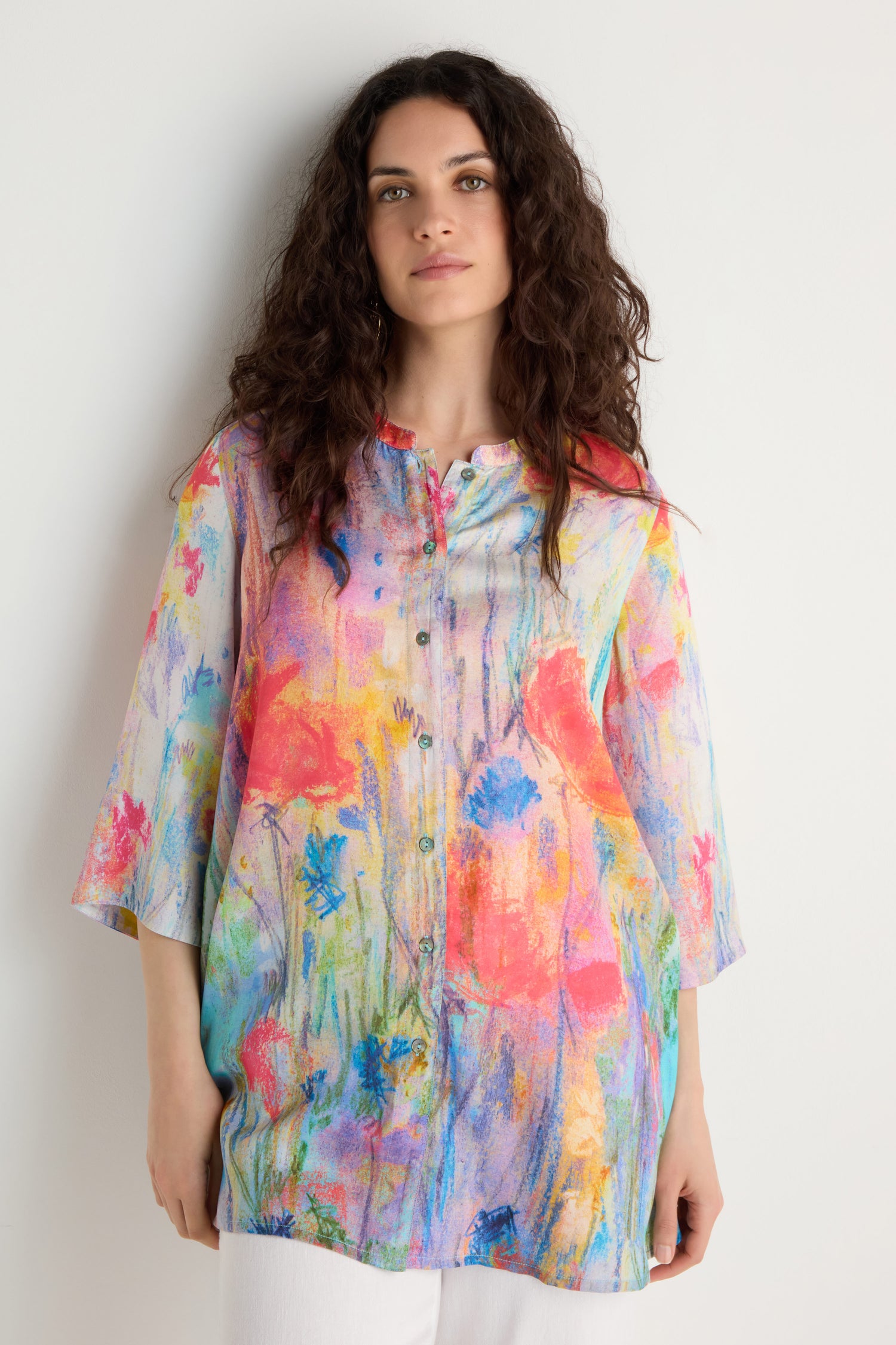 A woman stands against a white background wearing the Meadow Dreams Flared Shirt, featuring a colorful abstract floral print, paired with white pants.