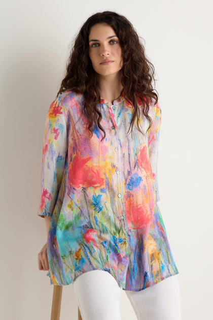 A woman with long curly hair wears the Meadow Dreams Flared Shirt, a vibrant floral print button-up blouse, paired with white pants as she sits against a plain light background.