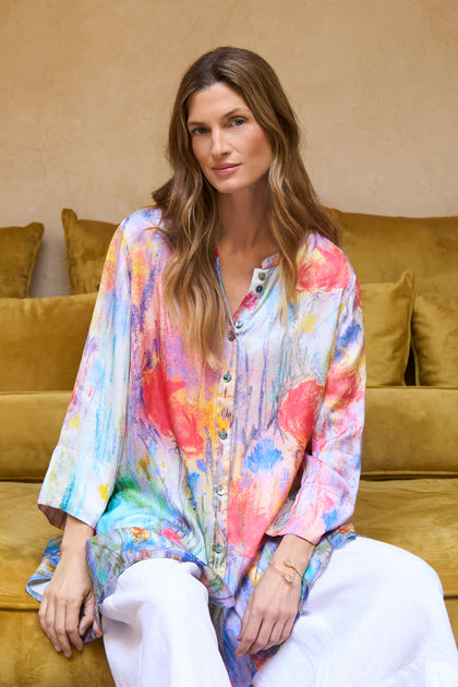 A woman with long brown hair sits on a yellow sofa, wearing the Meadow Dreams Flared Shirt and white pants, looking at the camera with a neutral expression.