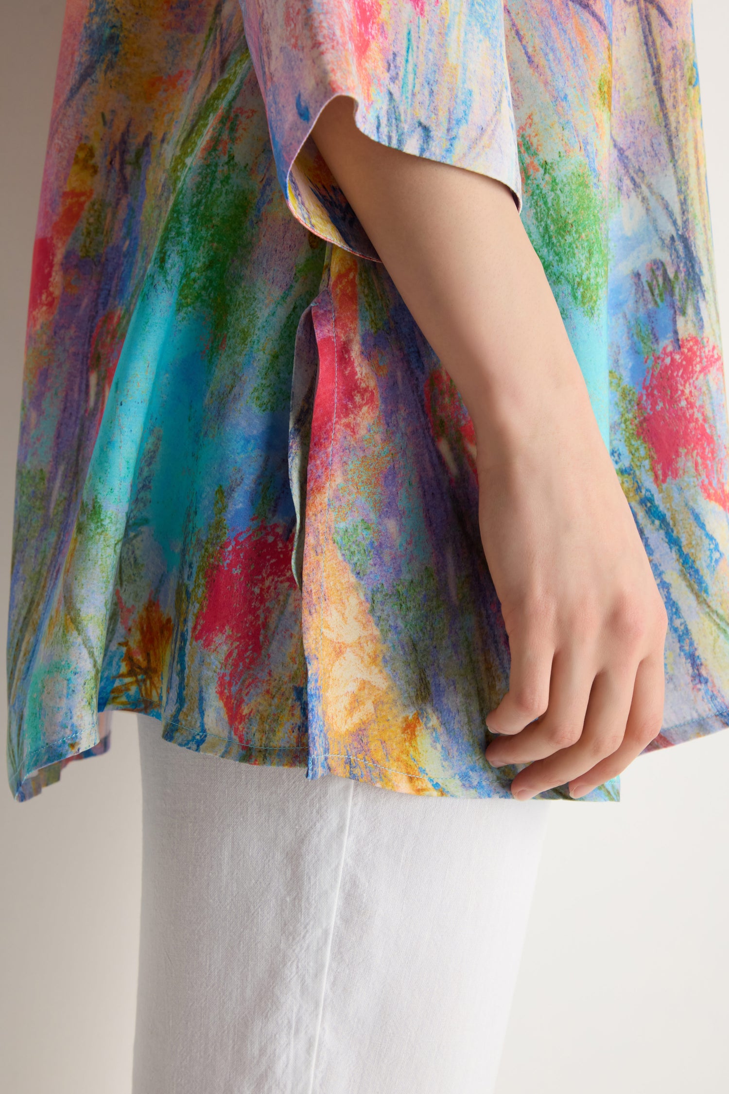 A person wears the Meadow Dreams Flared Shirt, featuring a vibrant abstract pattern and flared design, paired with white pants, their arm relaxed by their side.
