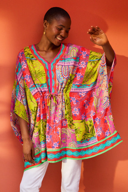 A person smiles while wearing a vibrant, patterned tunic with pink, green, and blue designs. They stand against an orange background, raising one arm. The luxurious bohemian vibe is unmistakable in their Tropical Patchwork Kaftan.