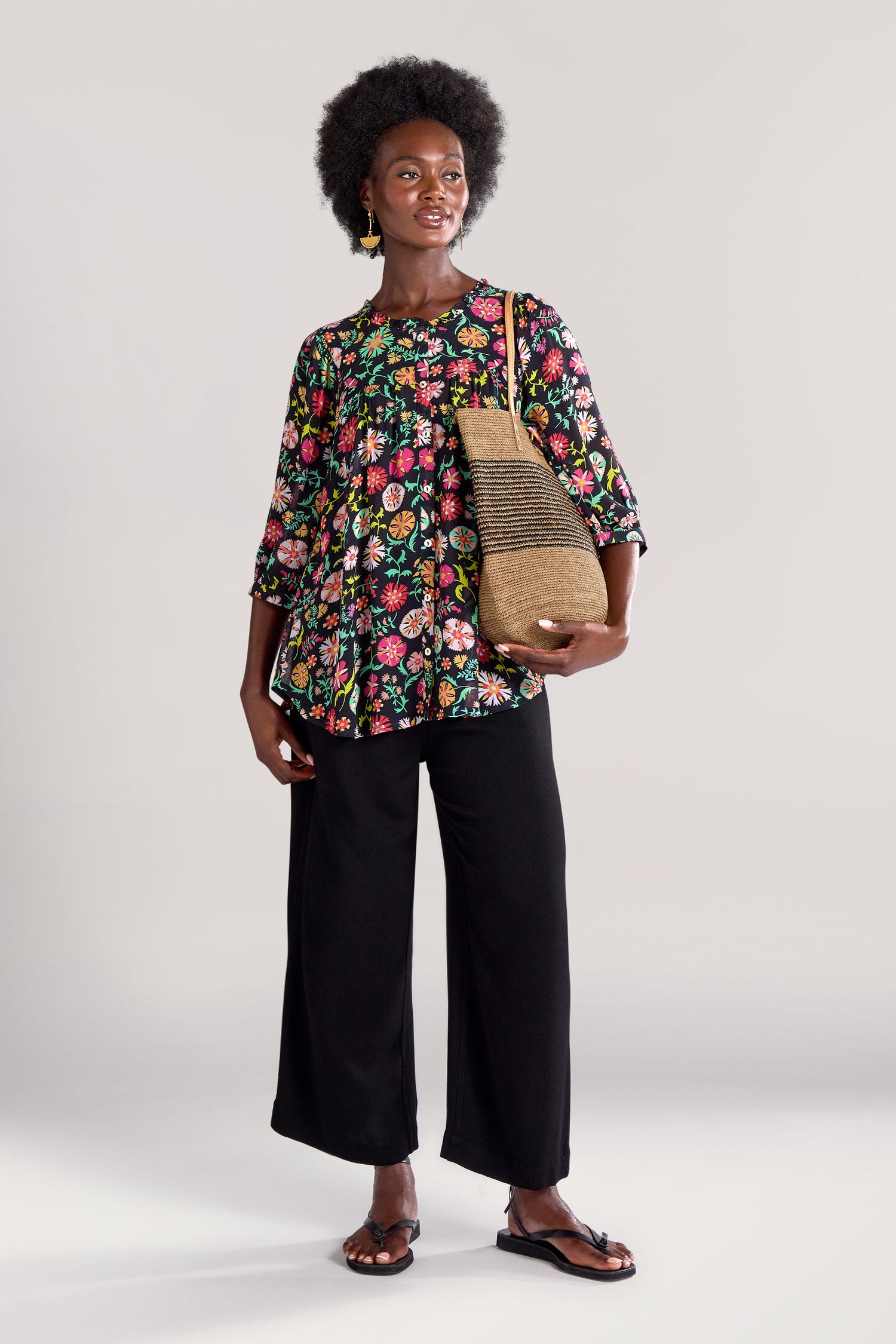 A person is standing against a plain background, dressed in a Folkloric Floral Shirt, black wide-leg pants, GRT5891-FFA sandals, and holding a straw handbag.