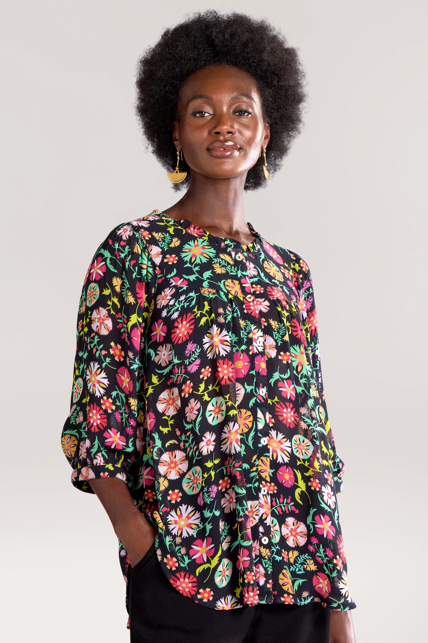 A person stands against a plain background, wearing a colorful floral blouse and black pants with hands in pockets, embodying the unique style of the Folkloric Floral Shirt.