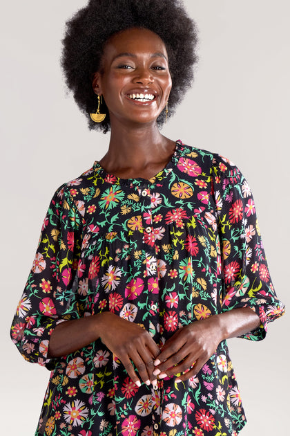 A person wearing a black floral-patterned blouse smiles while standing against a plain background, identified as Folkloric Floral Shirt.