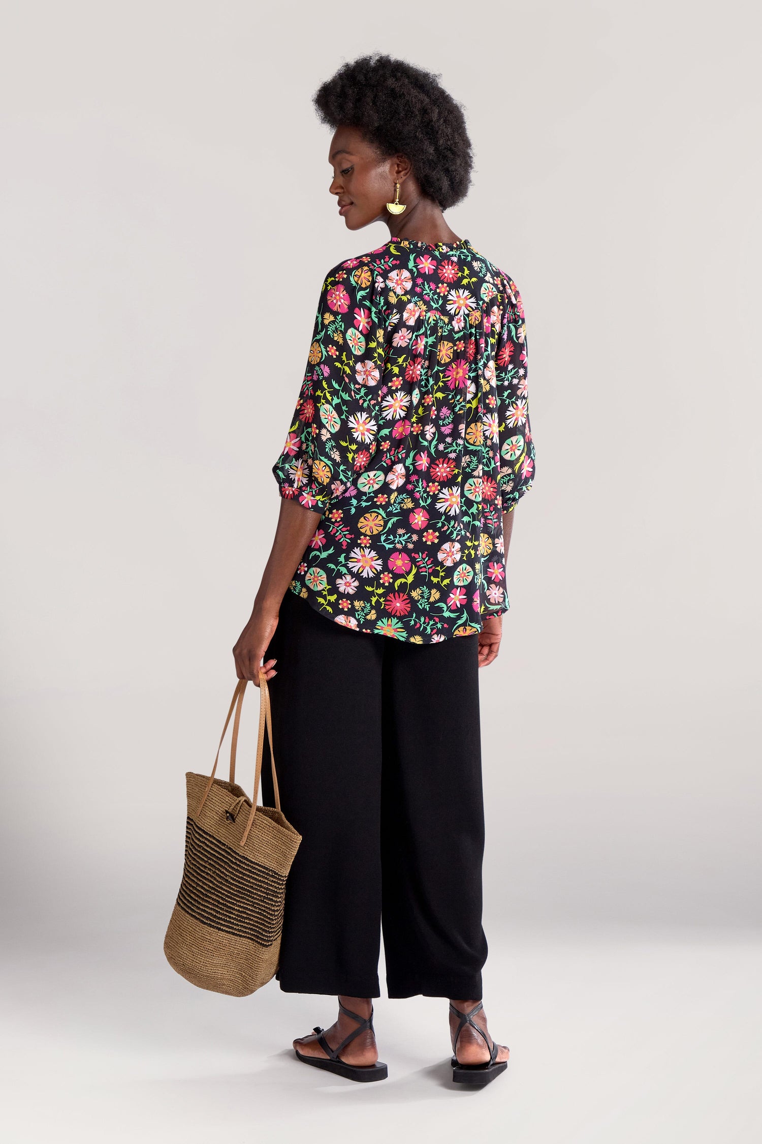 A person wearing a Folkloric Floral Shirt, black wide-legged pants, and sandals stands holding a GRT5891-FFA straw bag with black stripes. The background is plain.