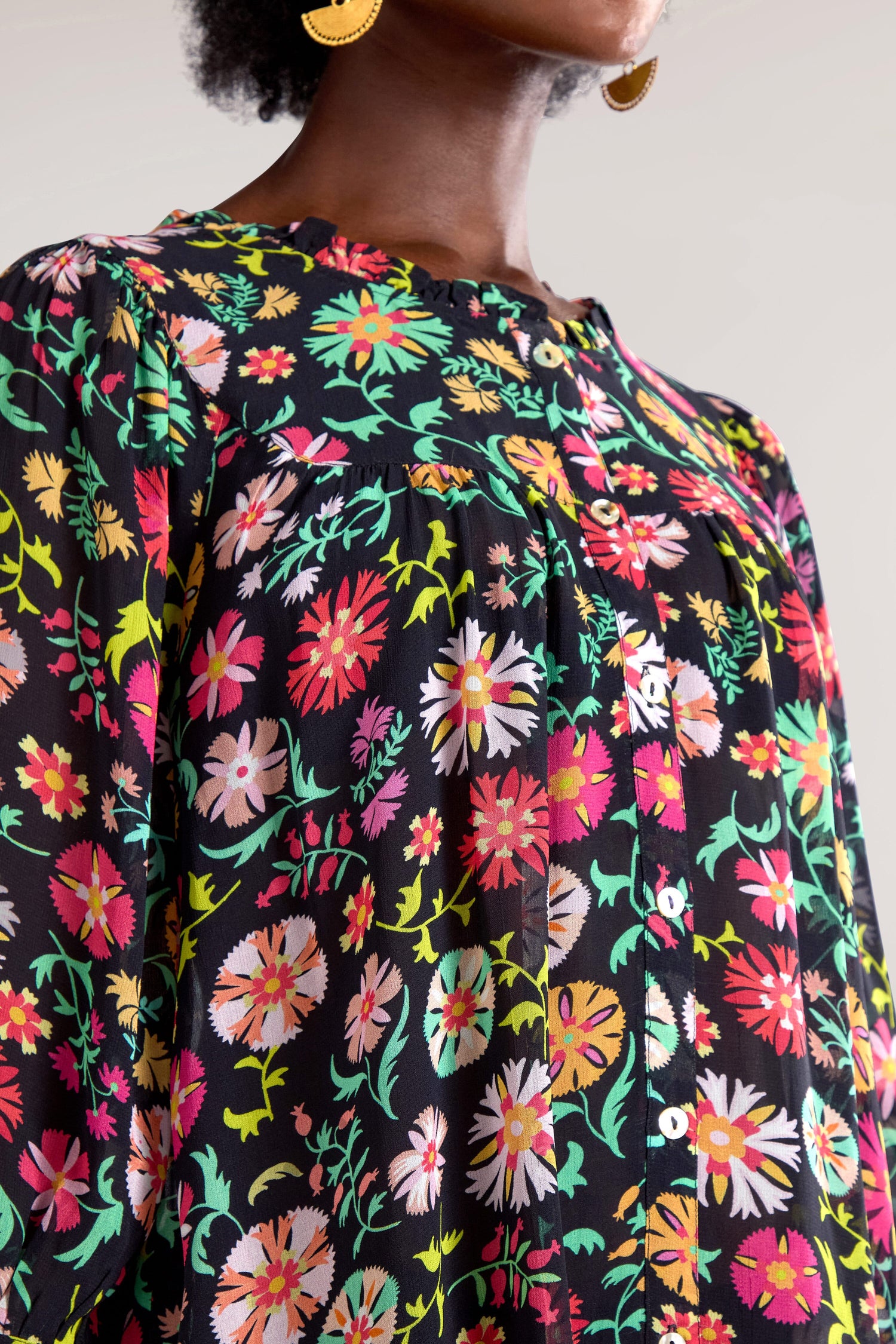 Close-up of a person wearing a black floral blouse with bright and colorful flower patterns. The person also wears large gold hoop earrings, resembling the stylish look of the Folkloric Floral Shirt.