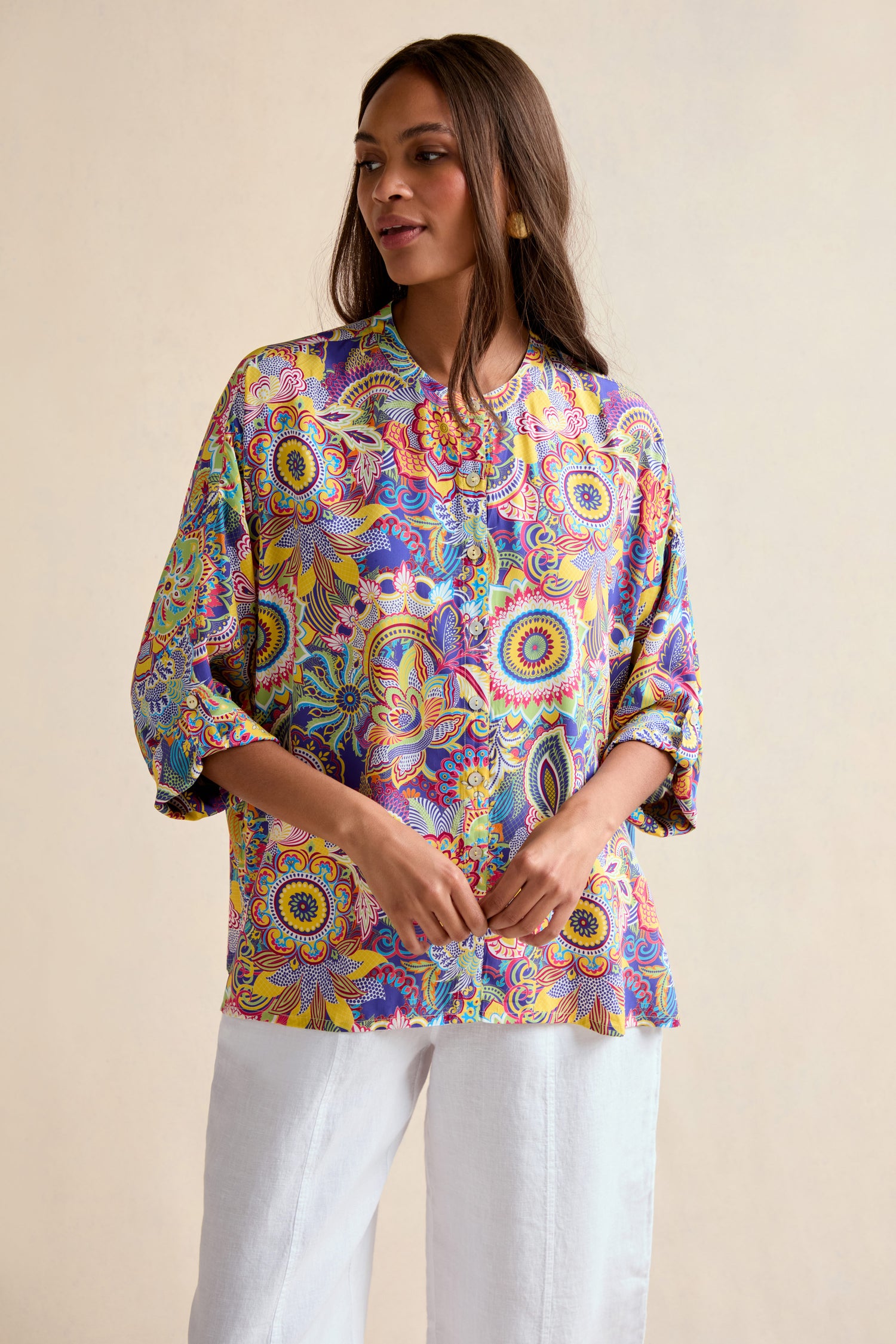 A woman wears the Summer Sarasa Relaxed Shirt, an oversized, colorful blouse with a mandala pattern, paired with white pants and posed against a simple, light background.