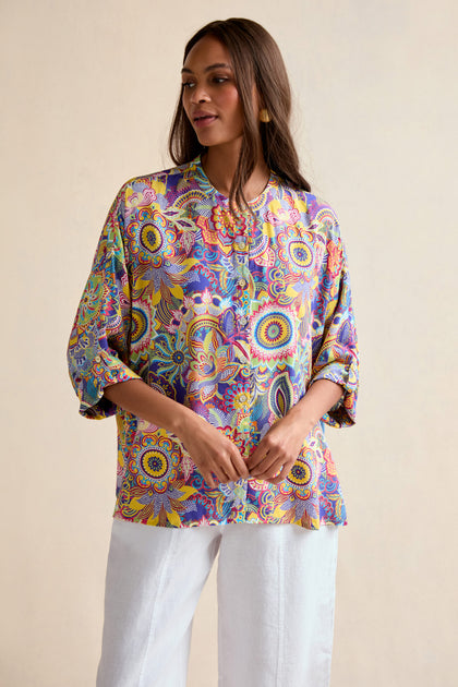 A woman wears the Summer Sarasa Relaxed Shirt, an oversized, colorful blouse with a mandala pattern, paired with white pants and posed against a simple, light background.