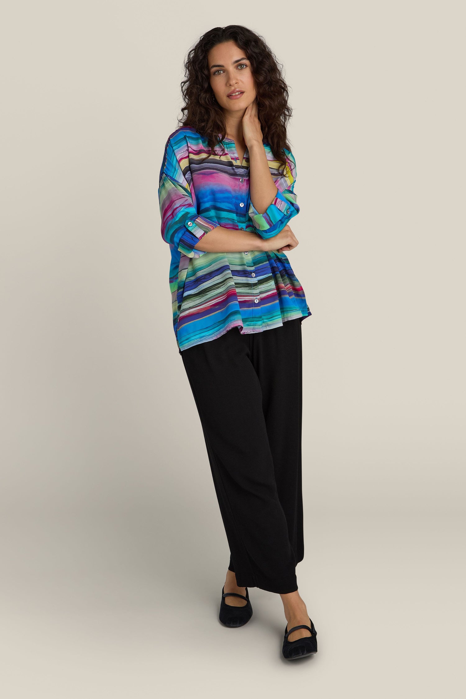 A person stands against a plain background, wearing the Watercolour Landscape Boxy Shirt made of viscose fabric, paired with black pants and black shoes. They have one hand touching their face and the other arm resting at their side.