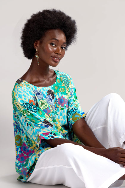 A person with an afro hairstyle wearing a brightly patterned Garden Spot Tunic and white pants is sitting on the floor, looking slightly to the side and smiling.