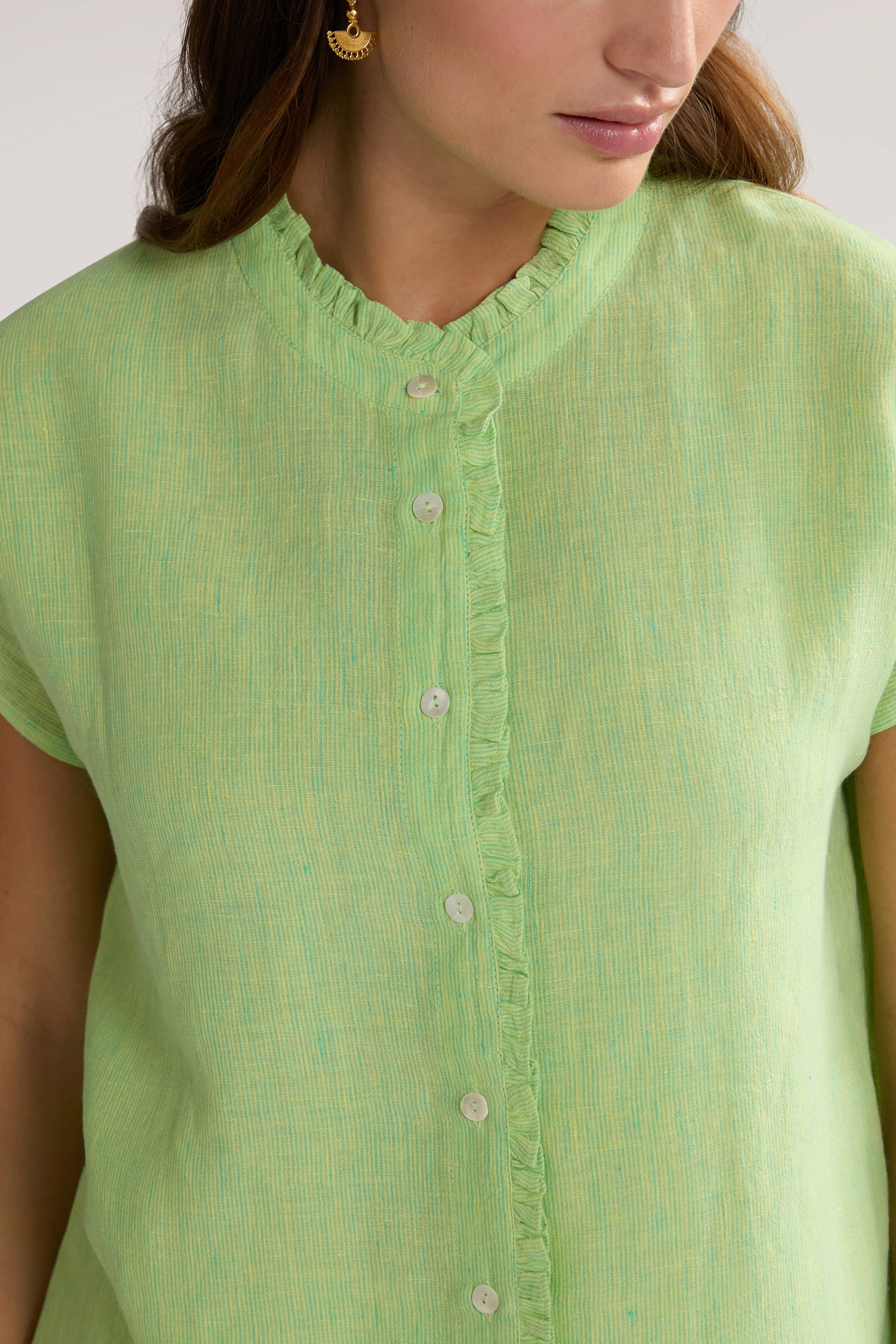 A person wearing a Linen Ticking Stripe Ruffle Front Shirt in light green with a ruffled collar detail is partially visible, showing only the upper torso and part of the face. They have long, brown hair and are wearing an earring.