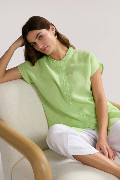 A woman in a Linen Ticking Stripe Ruffle Front Shirt and white pants sits on a beige armchair, leaning her head on her hand with a neutral expression.