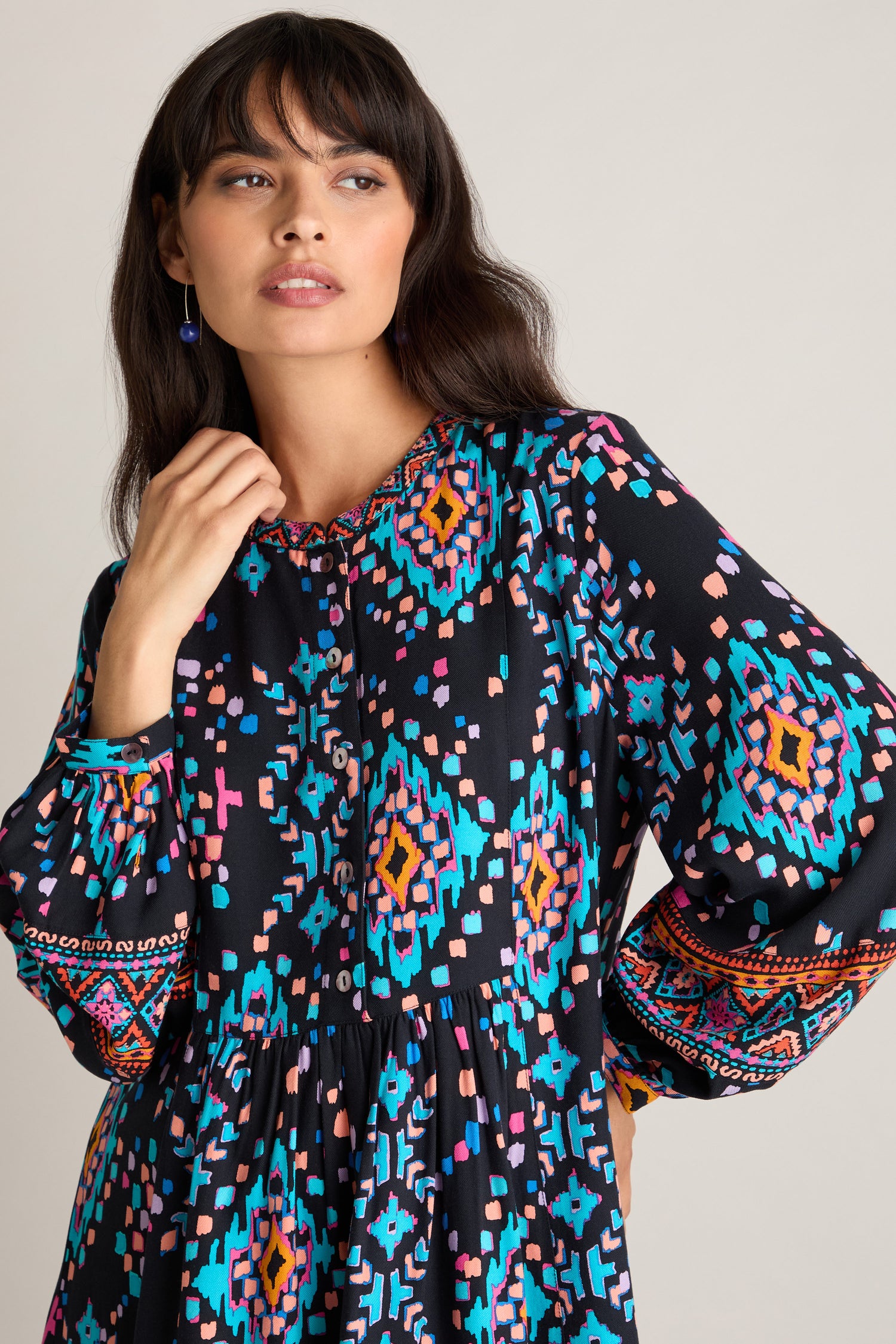 A woman with dark hair wears a vibrant Geo Kilim Tunic, glancing to the side with one hand touching her face.