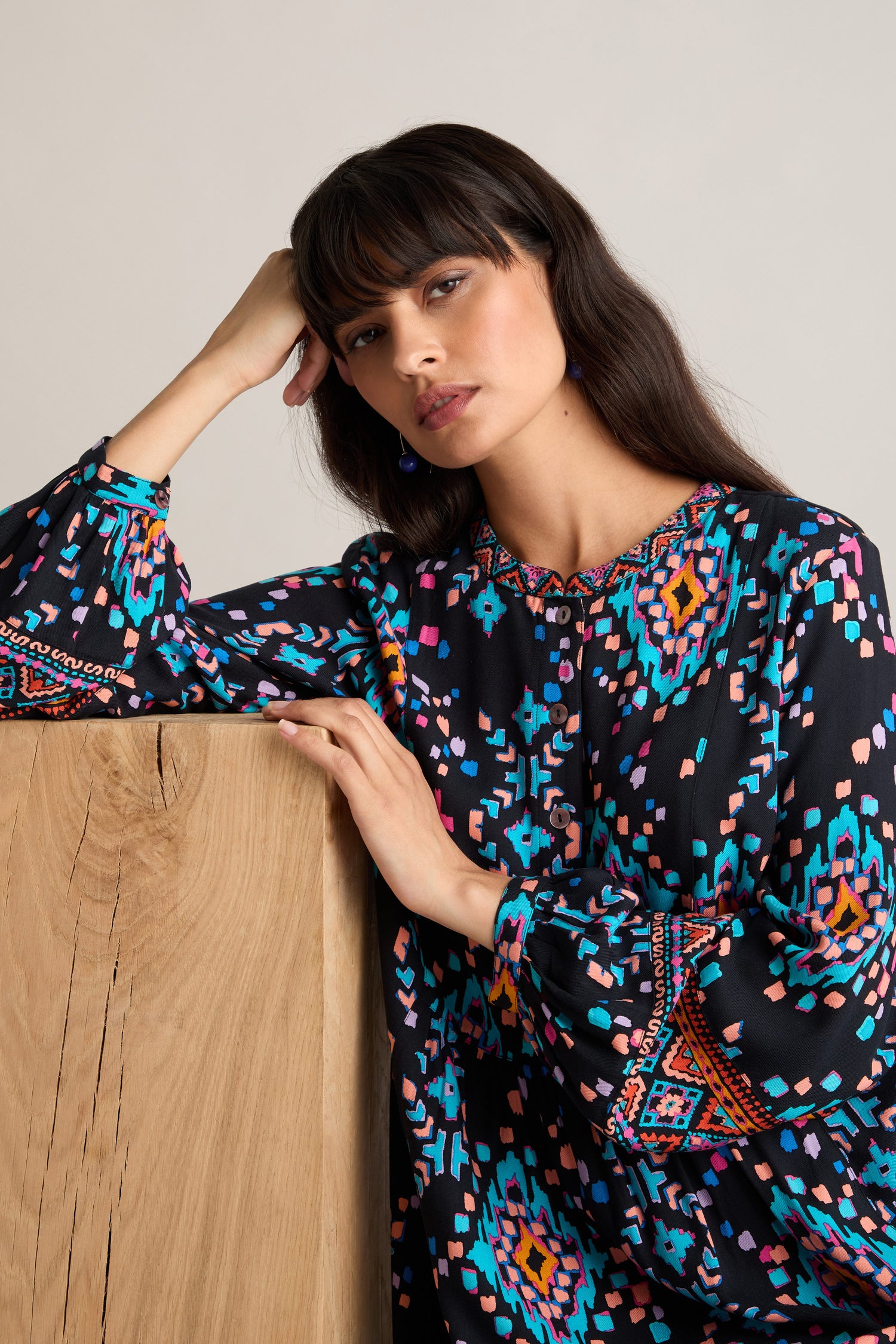 A woman with long dark hair, dressed in a vibrant Geo Kilim Tunic, leans on a wooden surface with her head resting on her hand and gazes into the camera.