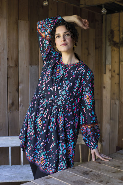 A person wearing the vibrant Geo Kilim Tunic stands inside, with one arm resting on a wooden surface and the other raised. The background shows wooden walls adorned with a hanging wreath.