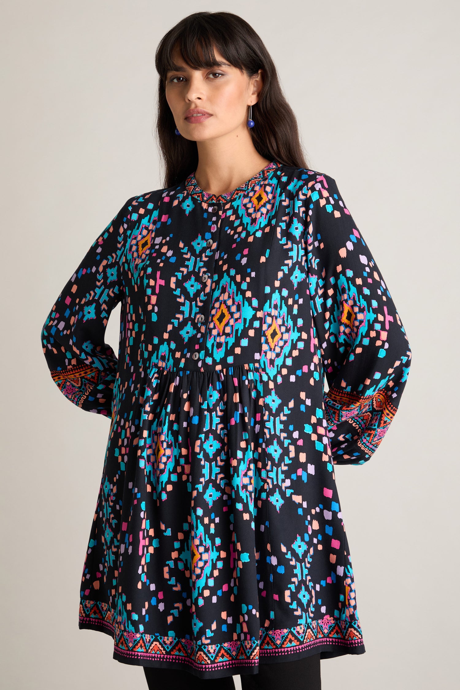 A person with long hair wears a Geo Kilim Tunic, which features a vibrant mix of blue, pink, and orange geometric designs on a black background and is accented by its stylish grandad collar.