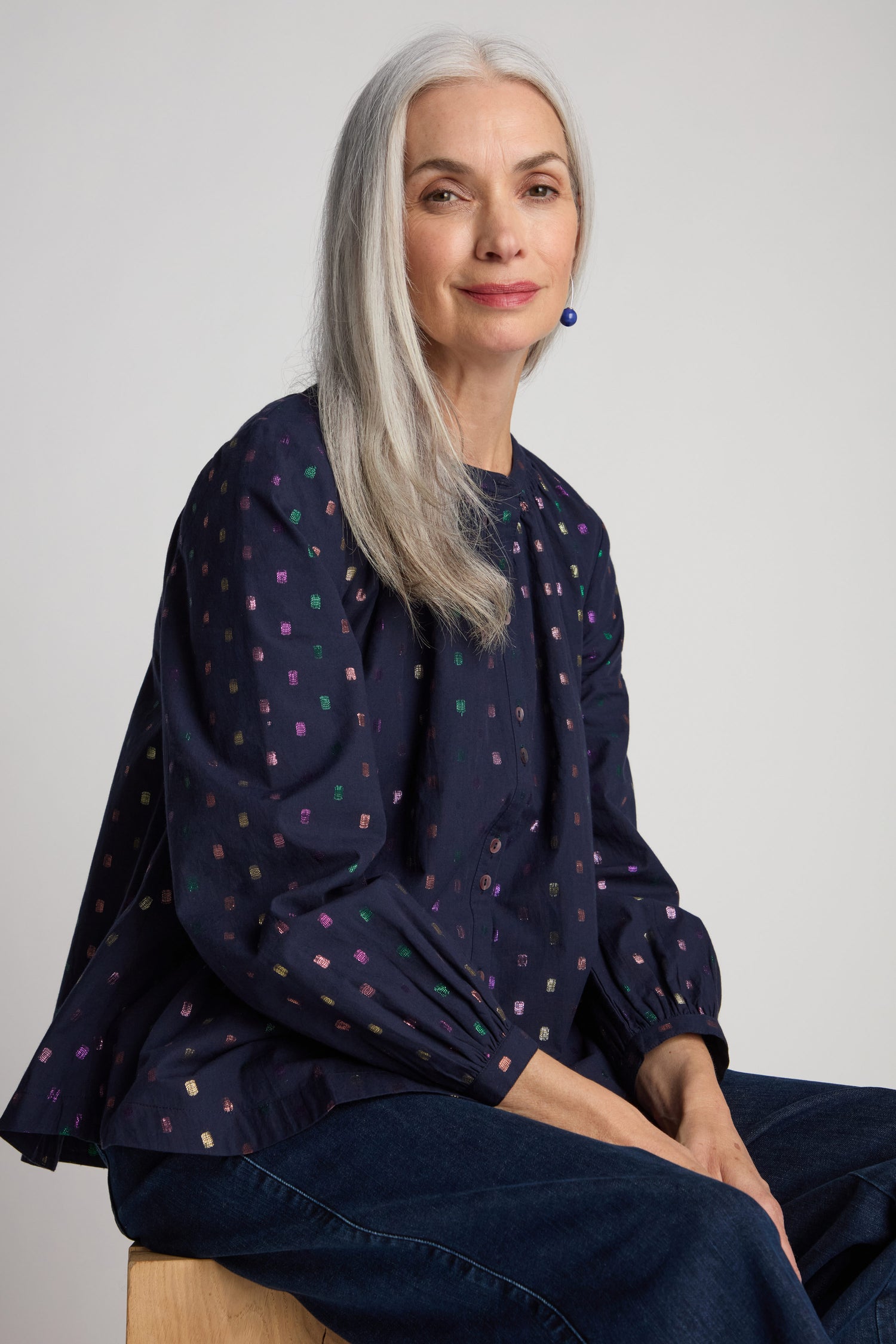 A woman with long gray hair, dressed in a Multi Square Lurex Shirt featuring shell buttons and paired with dark jeans, sits on a stool and looks at the camera.