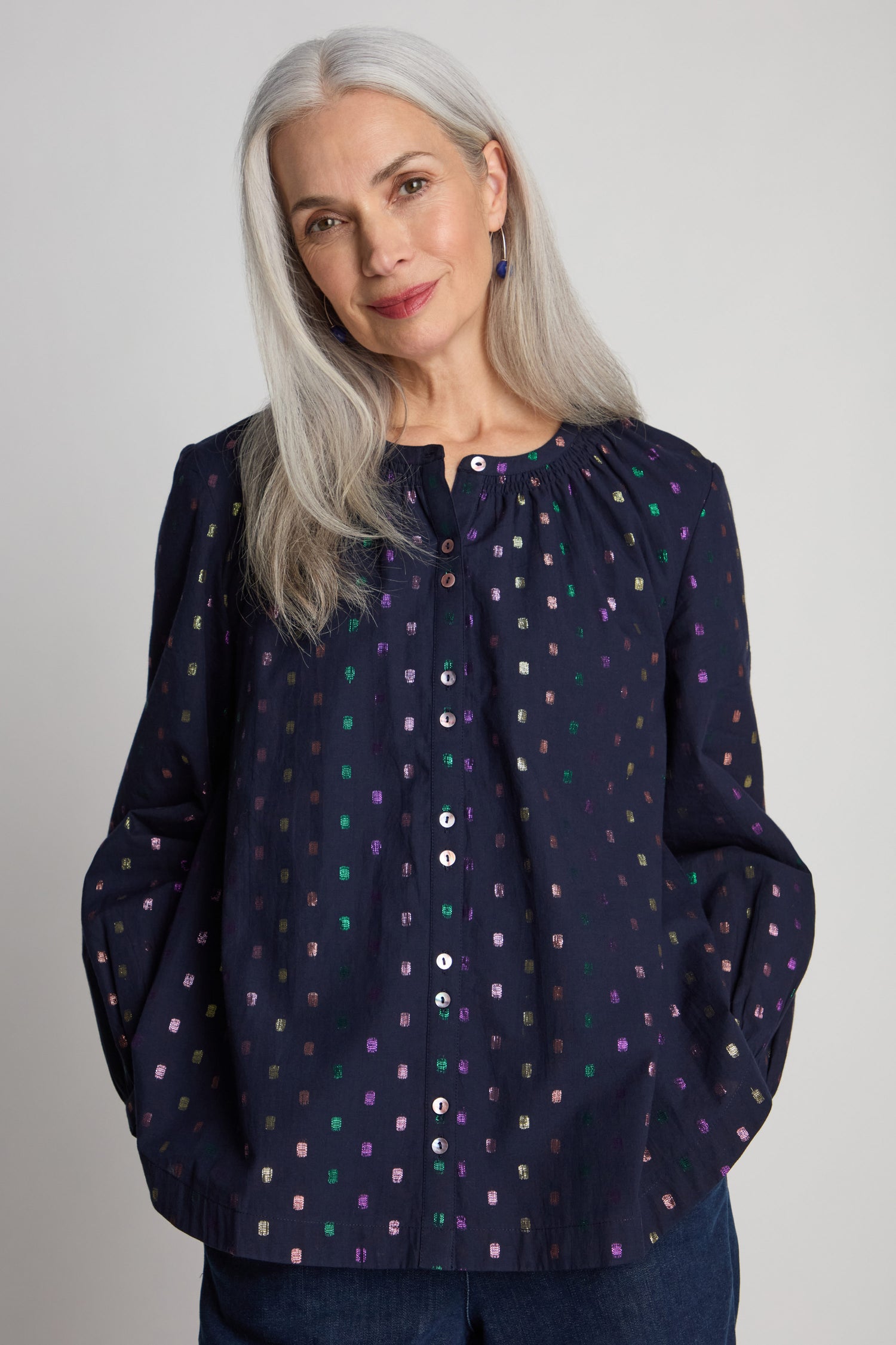 A woman with long gray hair, standing against a plain background, wears a navy Multi Square Lurex Shirt adorned with colorful square patterns and delicate shell buttons that add a subtle shine.