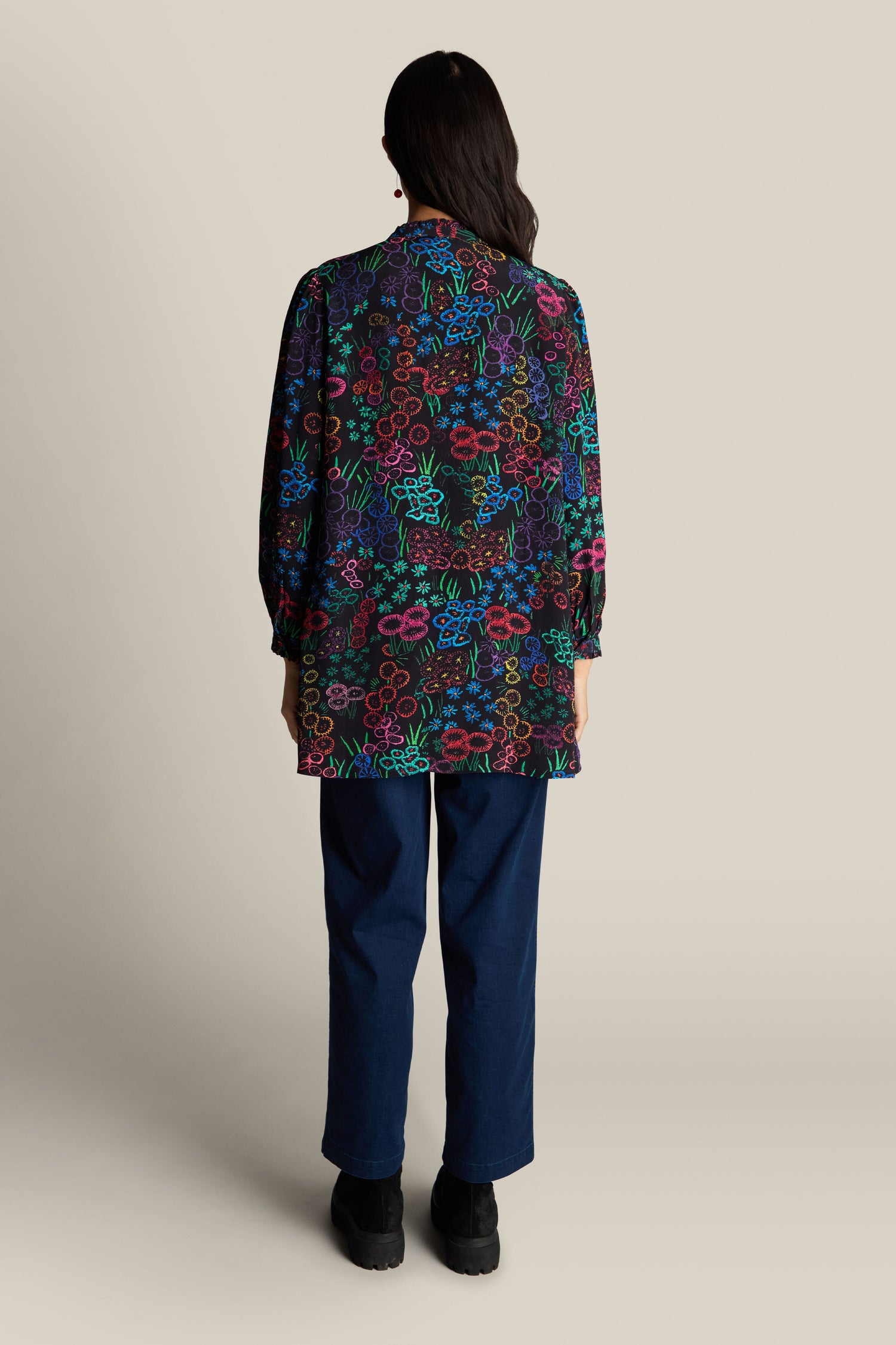 A person with long dark hair, dressed in the Embroidered Floral Print Tunic and dark blue pants, stands facing away against a neutral background.