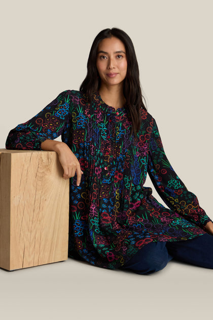 A woman with long dark hair, dressed in an Embroidered Floral Print Tunic, sits on the floor and leans against a wooden block, set against a beige background.