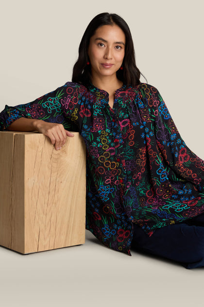 A person with long hair, wearing an "Embroidered Floral Print Shirt," sits on the floor leaning against a wooden block.