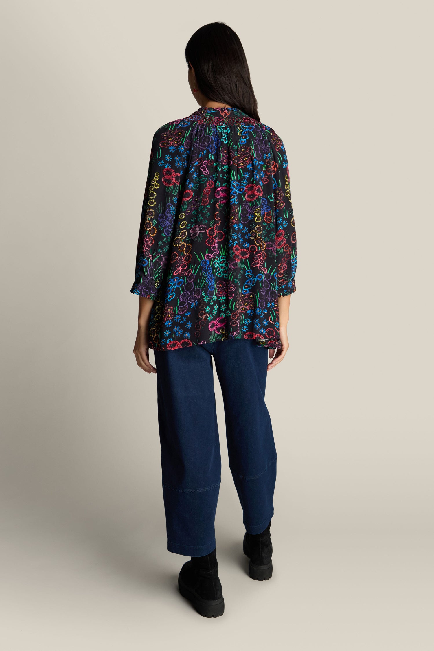 A person with long hair is seen from the back, wearing the Embroidered Floral Print Shirt featuring a colorful abstract pattern and dark blue pants, standing against a plain background.