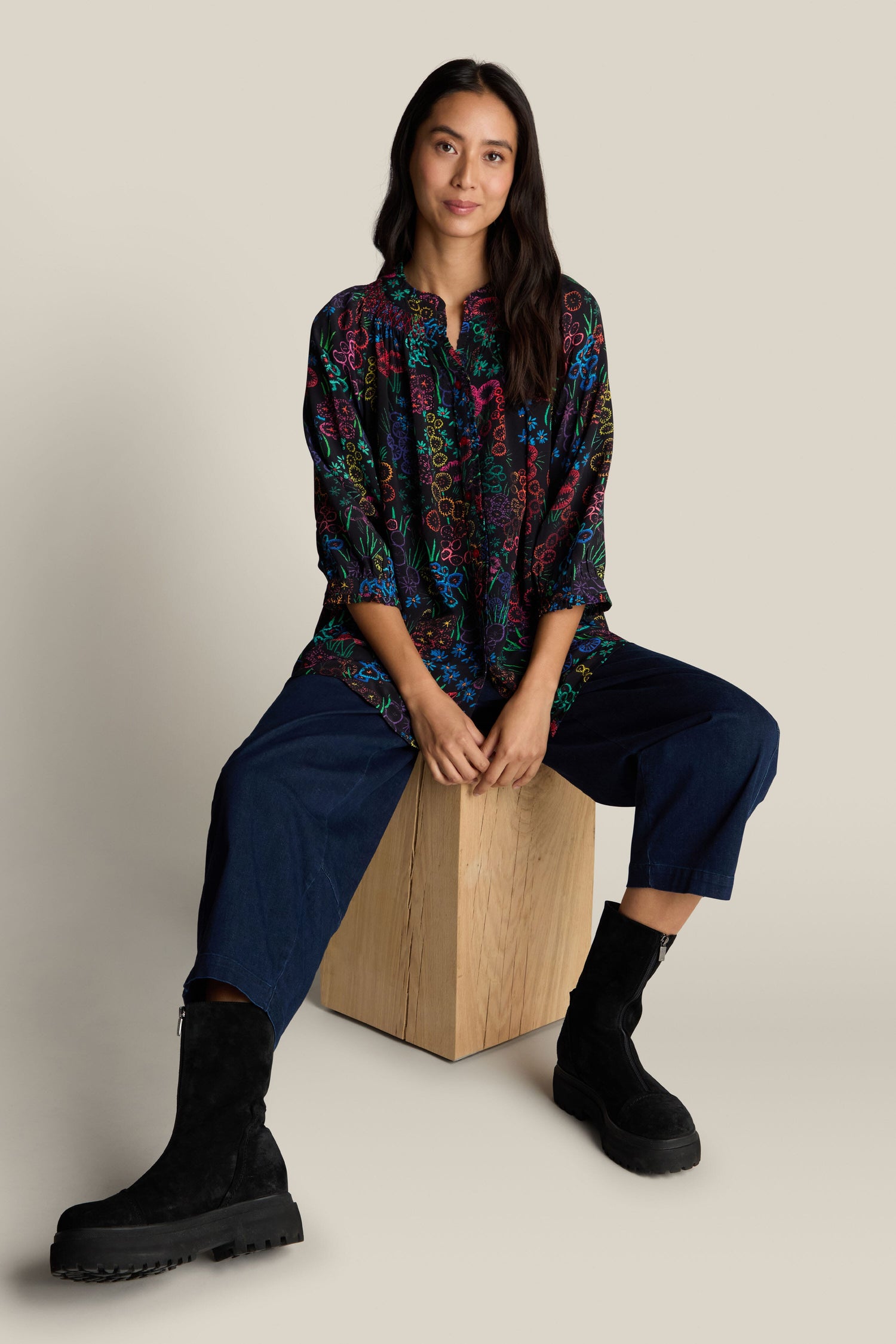 A person sits on a wooden block in a relaxed pose, wearing the Embroidered Floral Print Shirt with 3/4 length sleeves, blue pants, and black boots against a beige background.