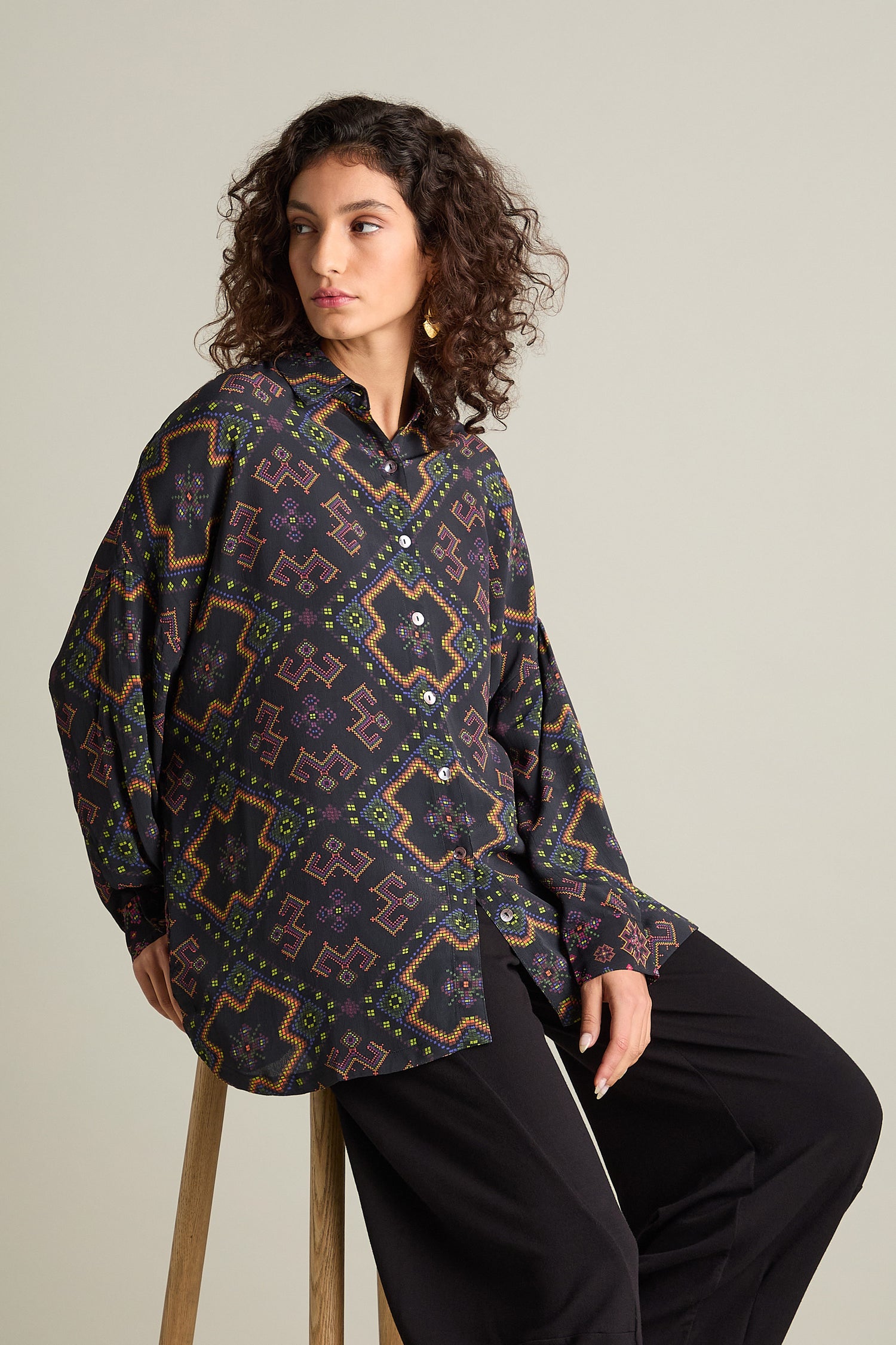 A person with curly hair, dressed in a Pixelated Jacquard Print Shirt featuring an oversized boxy fit and black pants, is seated on a wooden stool against a neutral background.