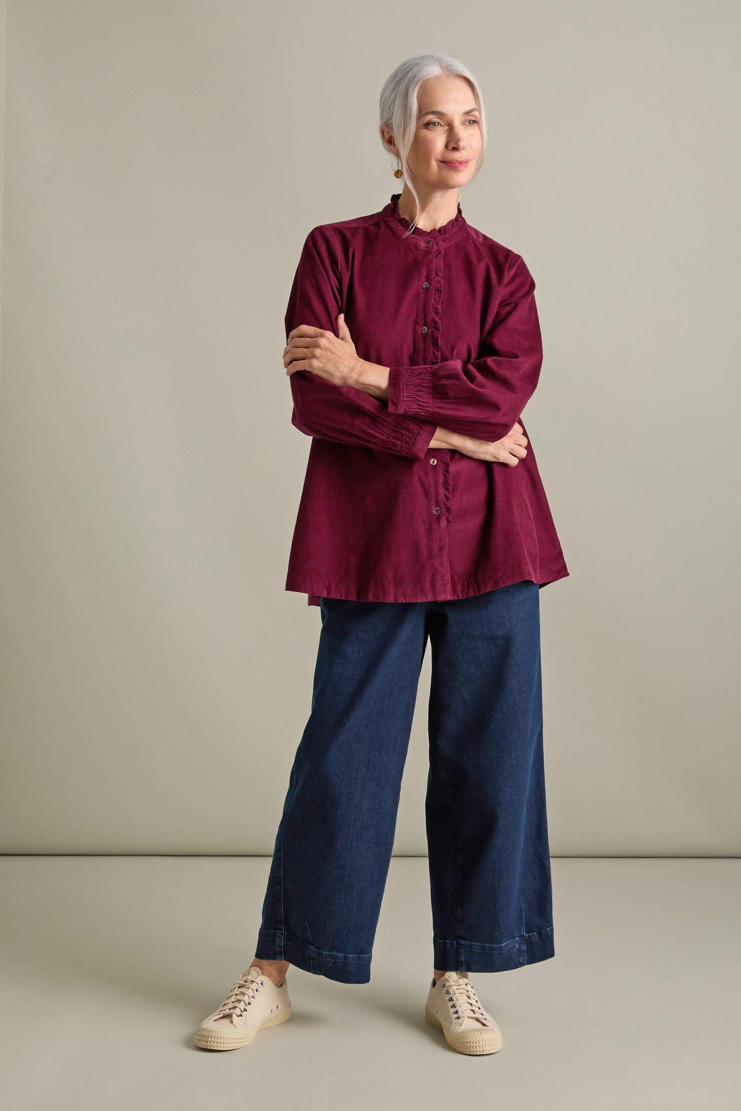 An older woman with gray hair stands confidently, wearing a burgundy Pinwale Cord Pleat Frill Placket Shirt, wide-legged blue jeans, and cream sneakers against a plain background.