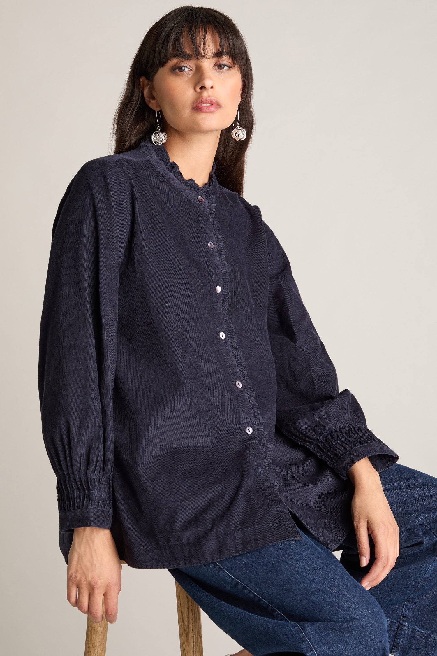 A person with long hair and fringe, wearing a Pinwale Cord Pleat Frill Placket Shirt made of luxurious cotton pincord and jeans, sits on a stool against a plain background.