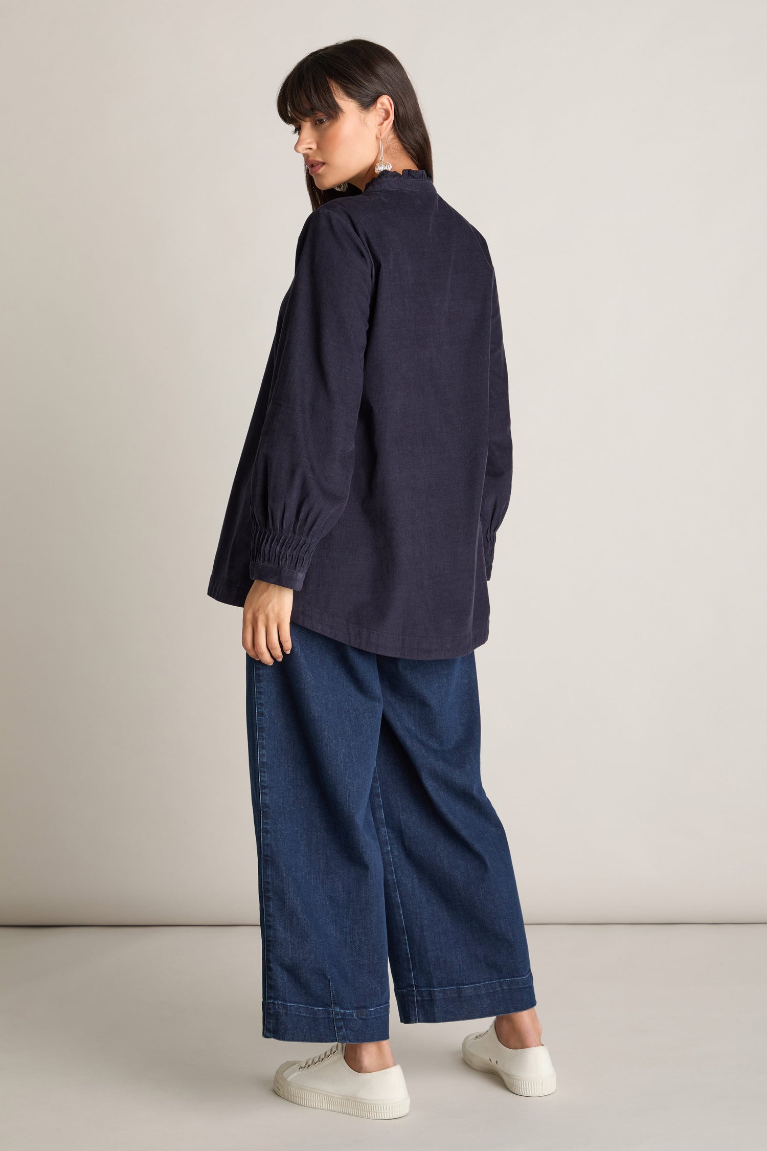 A woman with dark hair is wearing a Pinwale Cord Pleat Frill Placket Shirt, wide-leg jeans, and white sneakers. She stands facing slightly to the side, looking over her shoulder against a neutral background.