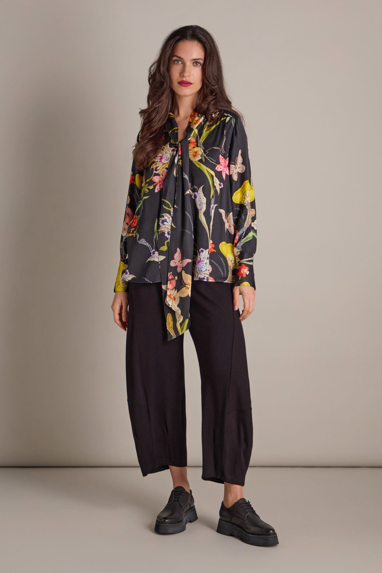 A woman in a Vintage Floral Tie Front Shirt paired with black pants stands confidently against a neutral background, her style accentuated by hand-embellished embroidery that offers an elegant touch.