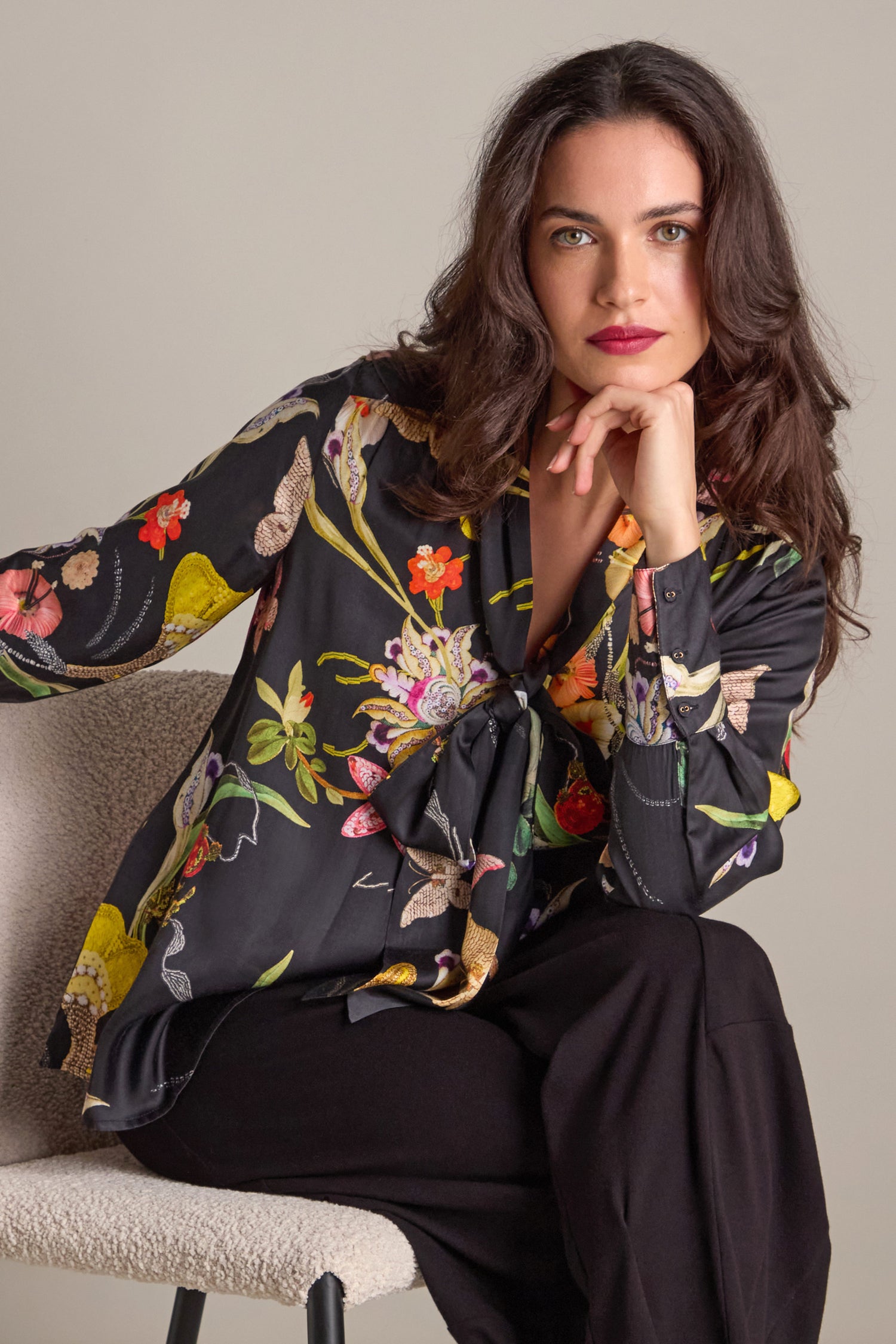 A woman with long dark hair sits on a chair, wearing a hand-embellished Vintage Floral Tie Front Shirt and black pants, with her chin resting on her hand.