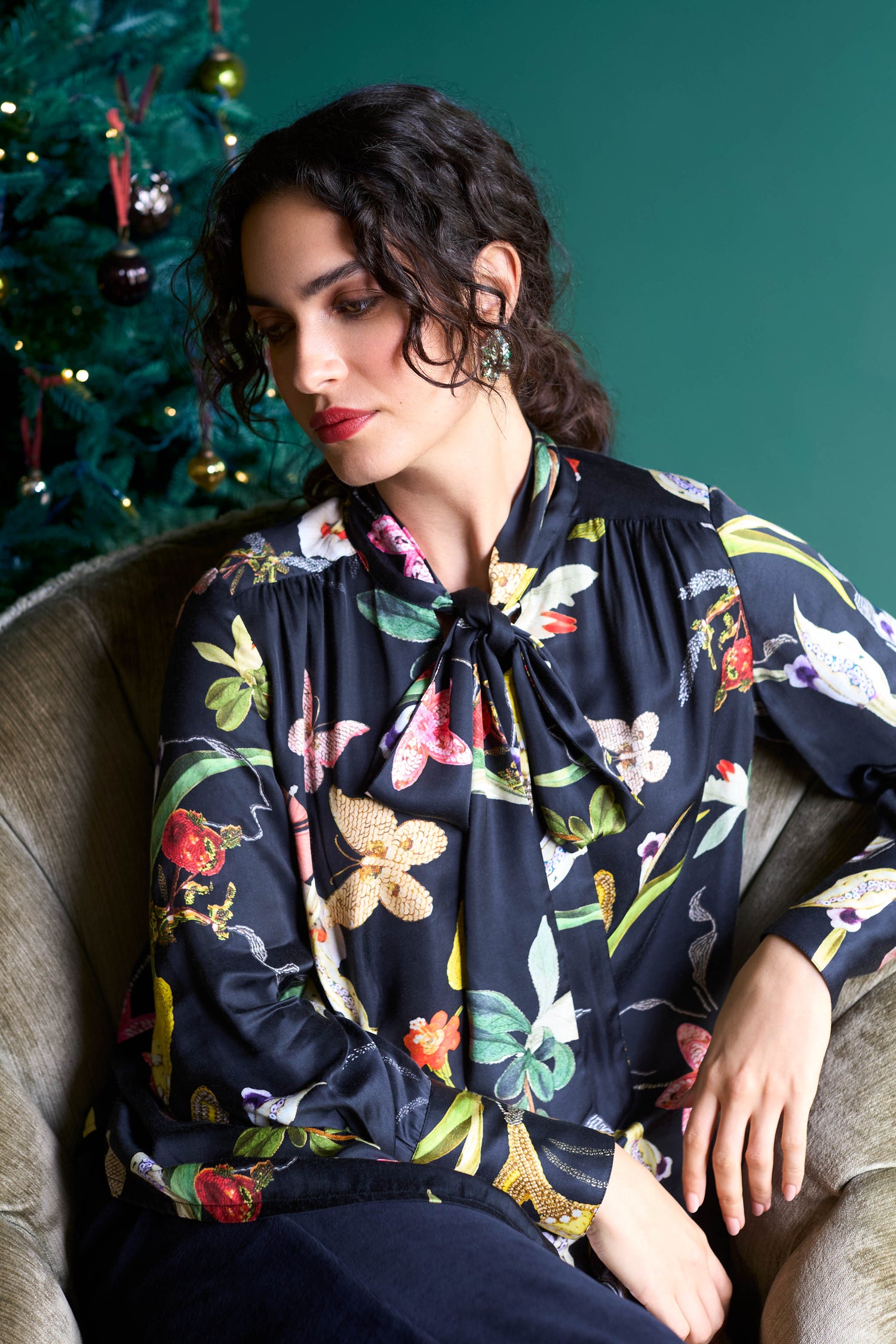 A woman with curly hair sits in a chair wearing a standout print Vintage Floral Tie Front Shirt. A decorated Christmas tree is visible in the background, adding to the festive charm.